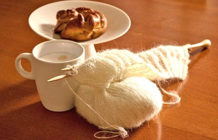 Cremefarbenes Garn mit Stricknadeln neben Tasse mit Schaum und Gebäck